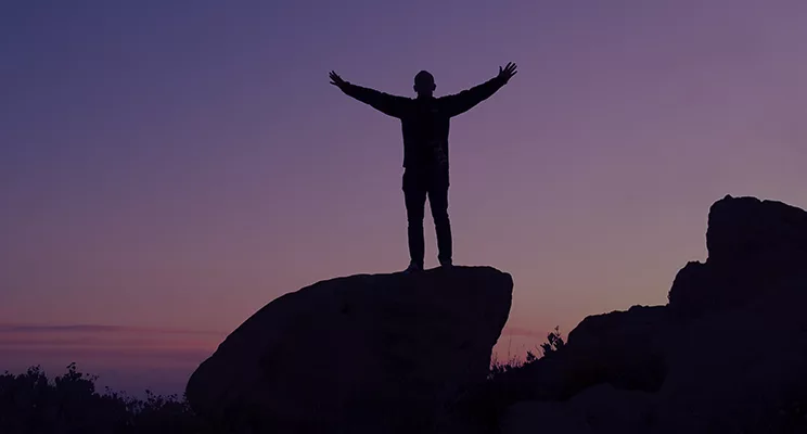 Silhouette of person with out stretched arms standing on a rock at dusk